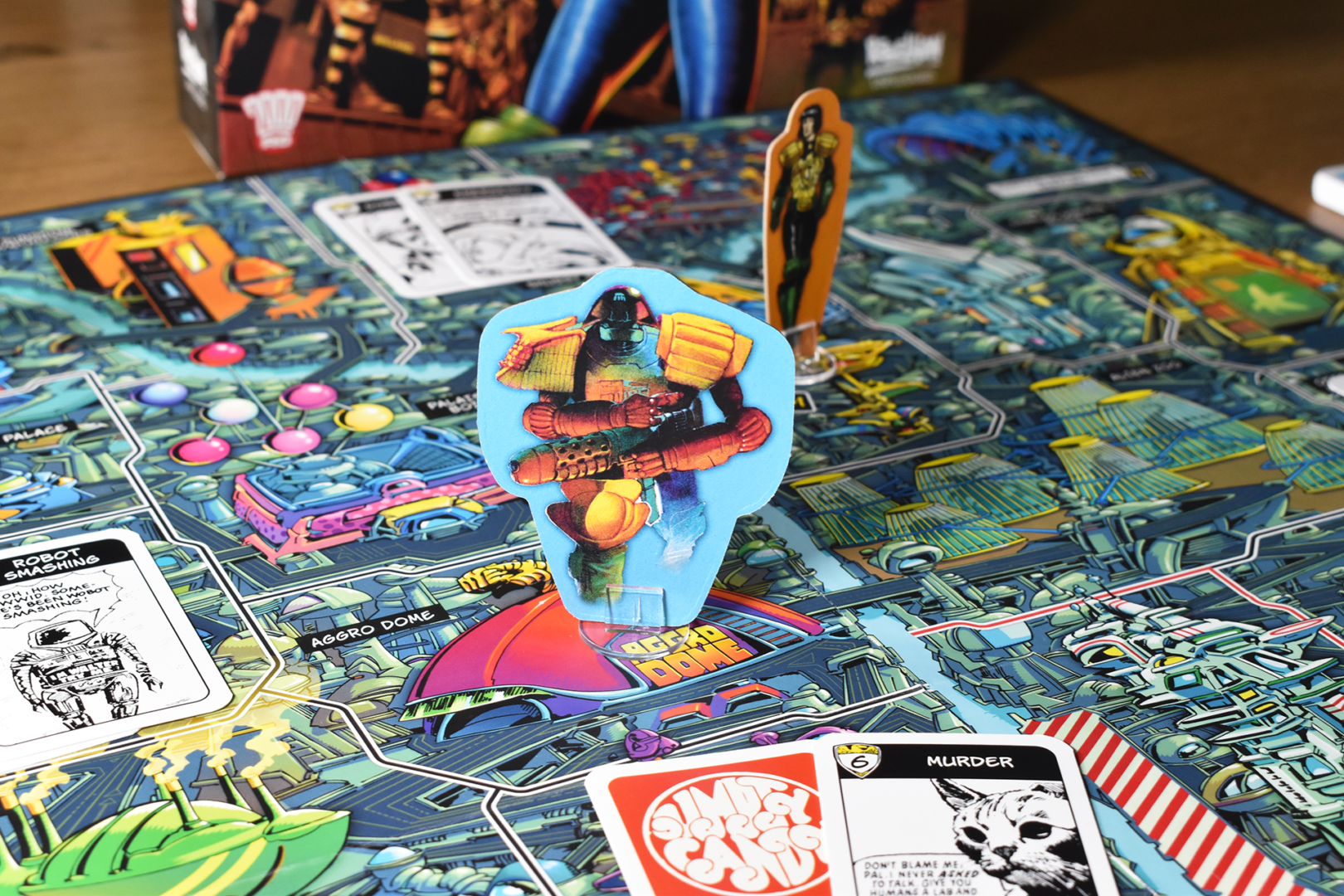 The Mechanismo standing on the board from Judge Dredd: The Game of Crime-Fighting in Mega-City One. In the background we can see another Judge, and Perp and Crime cards are placed around the board.