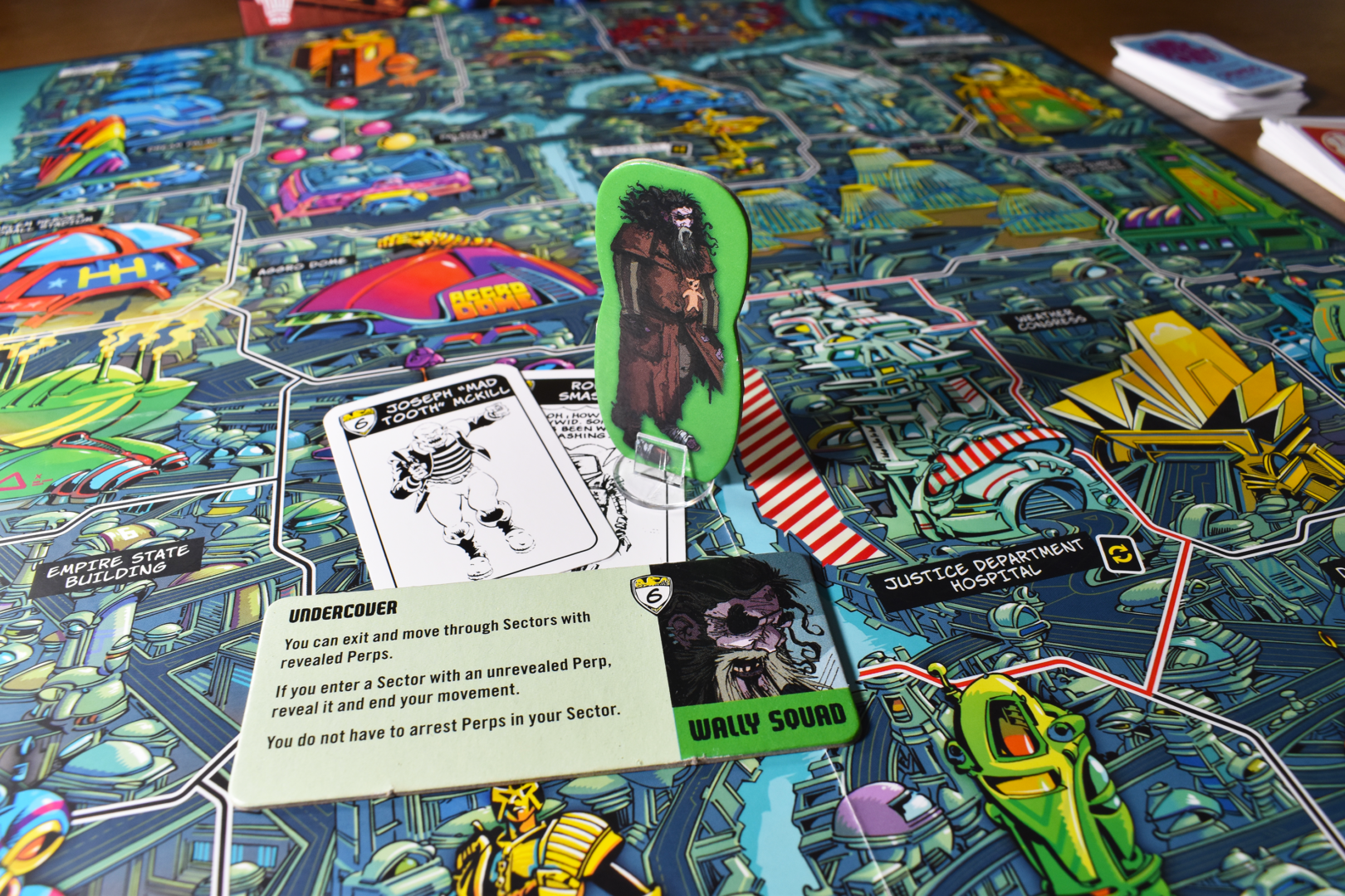 A standee of Dirty Frank standing a board depicting Mega-City One, with a Perp and Crime card beneath him and his Specialist Token in front of him.

The token reads:
"Undercover
You can exit and move through Sectors with revealed Perps.
If you enter a Sector with an unrevealed Perp, reveal it and end your movement.
You do not have to arrest Perps in your sector"