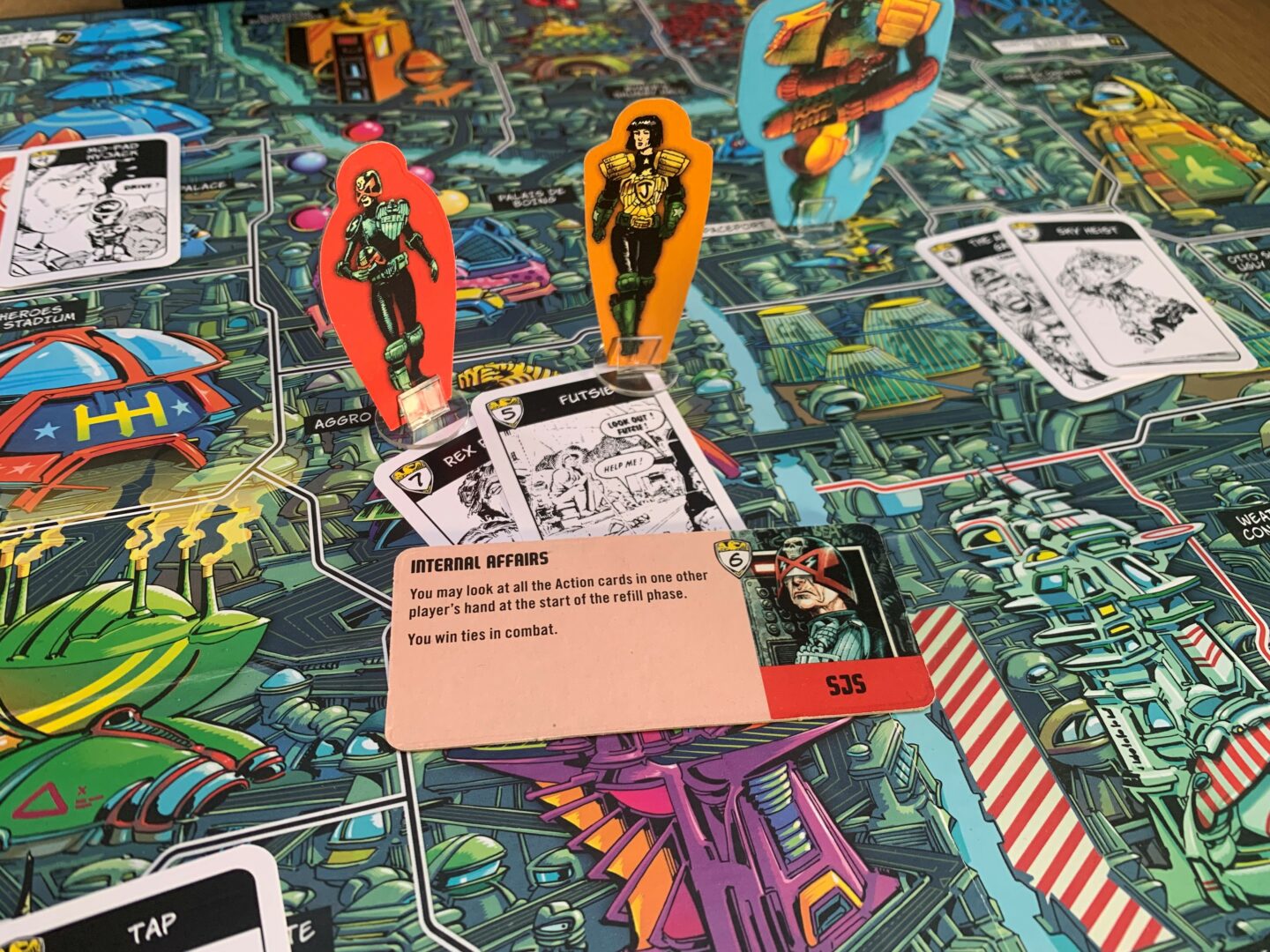 The SJS standee placed beside the Chief Judge standee on the Mega-City One map from Judge Dredd: The Game of Crime Fighting in Mega-City One.

In front of the Judges is a Crime and Perp card, as well as the Specialist Judge ability token that reads:
“Internal Affairs
You may look at all the Action cards in one other player’s hand at the start of the refill phase.
You win ties in combat.”
